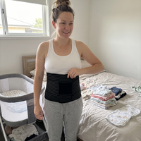 Woman a few weeks after birth wearing the black postpartum belly wrap over a white tank top in her bedroom